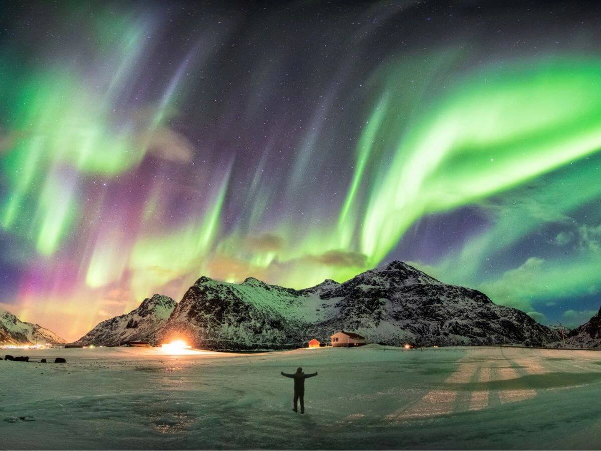 Un hombre de pie en la nieve, con los brazos extendidos, bajo una vibrante aurora boreal verde y púrpura en el cielo nocturno.