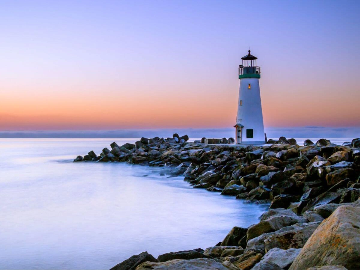 Un faro se alza sobre escarpadas rocas, iluminado por los cálidos tonos de un cielo al atardecer.