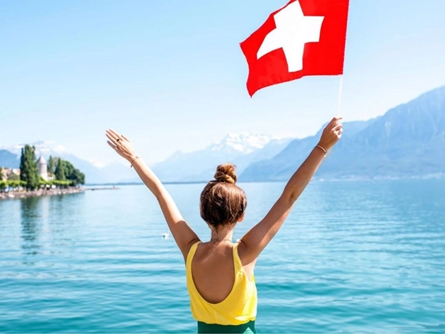 Mujer sosteniendo una bandera suiza frente a un lago, con un paisaje natural de fondo que refleja tranquilidad y belleza.