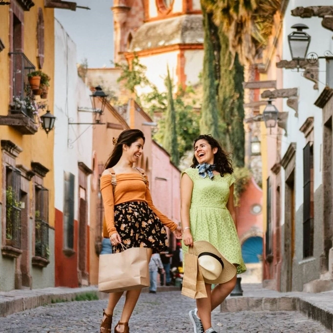 Dos mujeres caminan por una calle empedrada, sosteniendo bolsas de compras en sus manos.