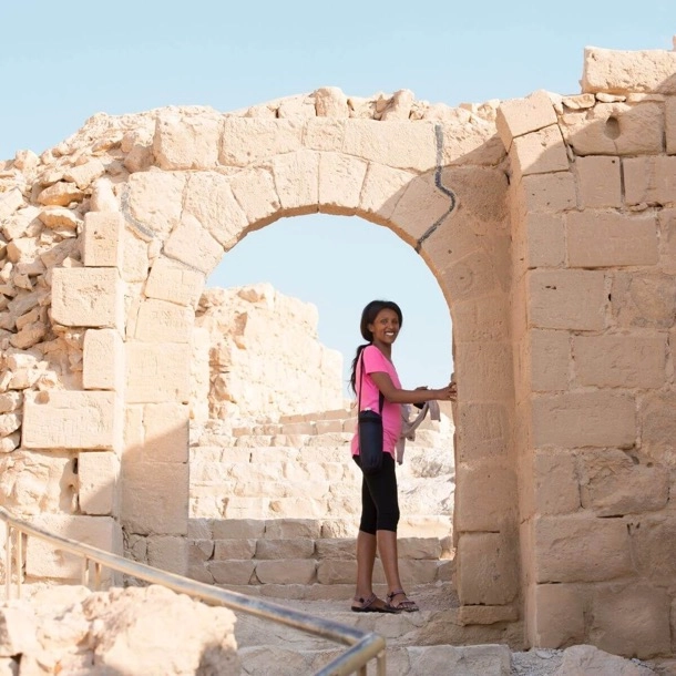 The image shows an individual standing in an ancient stone archway, which appears to be part of a historical site or ruins. The setting suggests a place of archaeological interest, possibly in a desert region given the clear skies and sandy terrain.