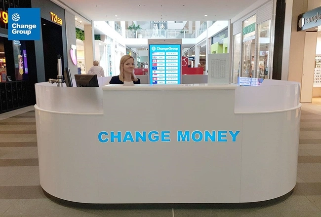 Una mujer está de pie detrás de un mostrador, mostrando un cartel de cambio de dinero, dispuesta a ayudar a los clientes con las transacciones.