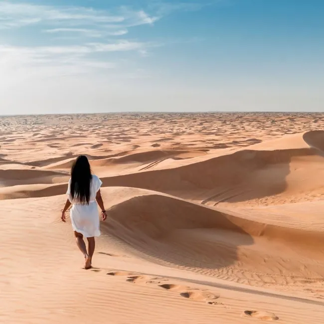 Una mujer camina sola en un vasto paisaje desértico, rodeada de dunas de arena bajo un cielo azul despejado.