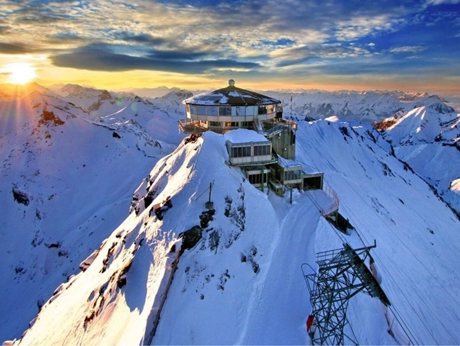 Cima de una montaña con un telesilla y una casa en la parte superior, rodeada de un paisaje nevado.