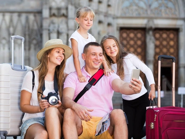 Una familia posa para un selfie, rodeada de equipaje y una cámara, captando juntos un alegre momento de viaje.