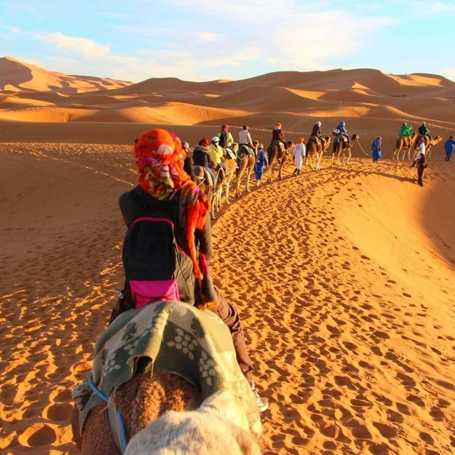 Caravana de camellos con jinetes en un paisaje de dunas desérticas al atardecer.