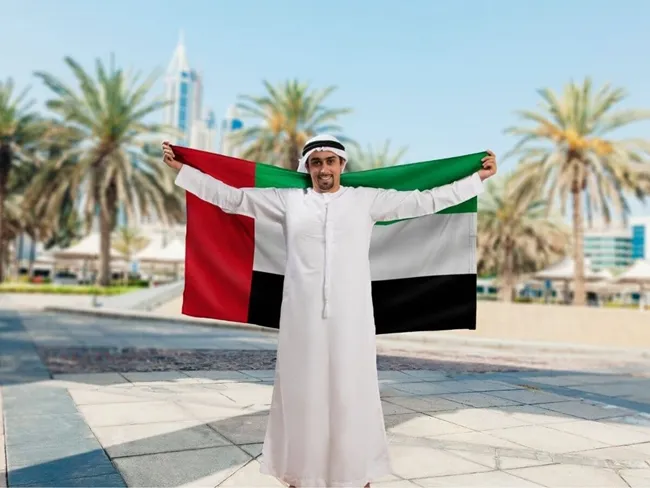 Un hombre sostiene con orgullo la bandera de los EAU con el vibrante horizonte de la ciudad como telón de fondo.